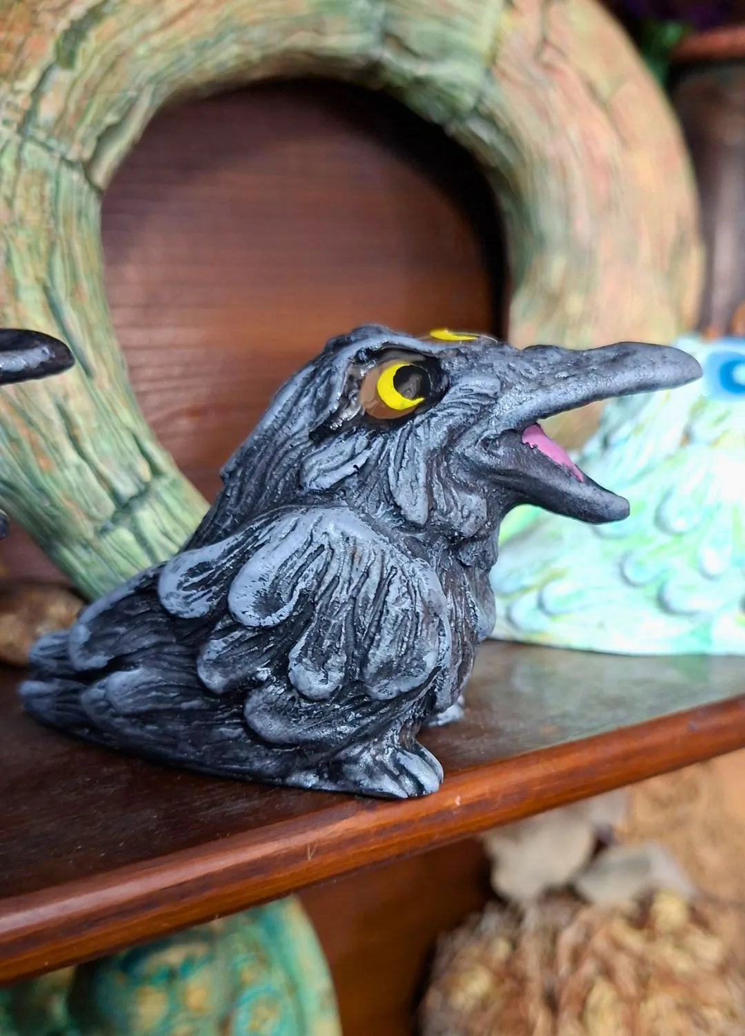 Close up of a handmade gothic raven statue in dark grey and black with detailed feathers and bright orange eyes, displayed on a wooden shelf.