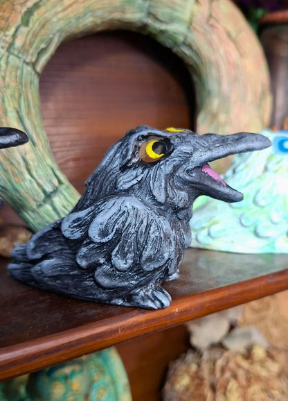 Close up of a handmade gothic raven statue in dark grey and black with detailed feathers and bright orange eyes, displayed on a wooden shelf.