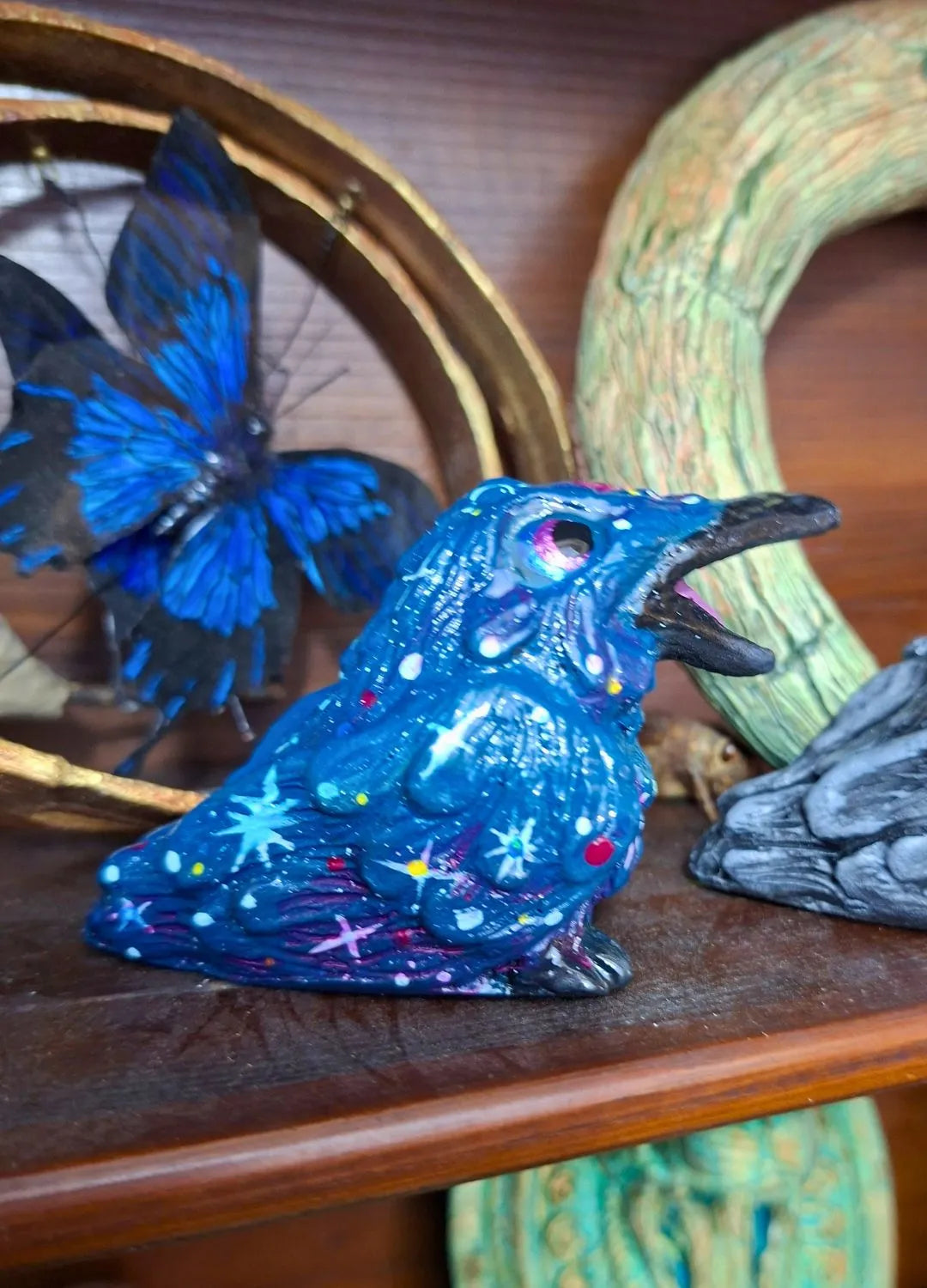 Detailed shot of a celestial blue resin raven statue painted with pink nebula clouds and tiny white stars, sitting next to a blue butterfly prop.