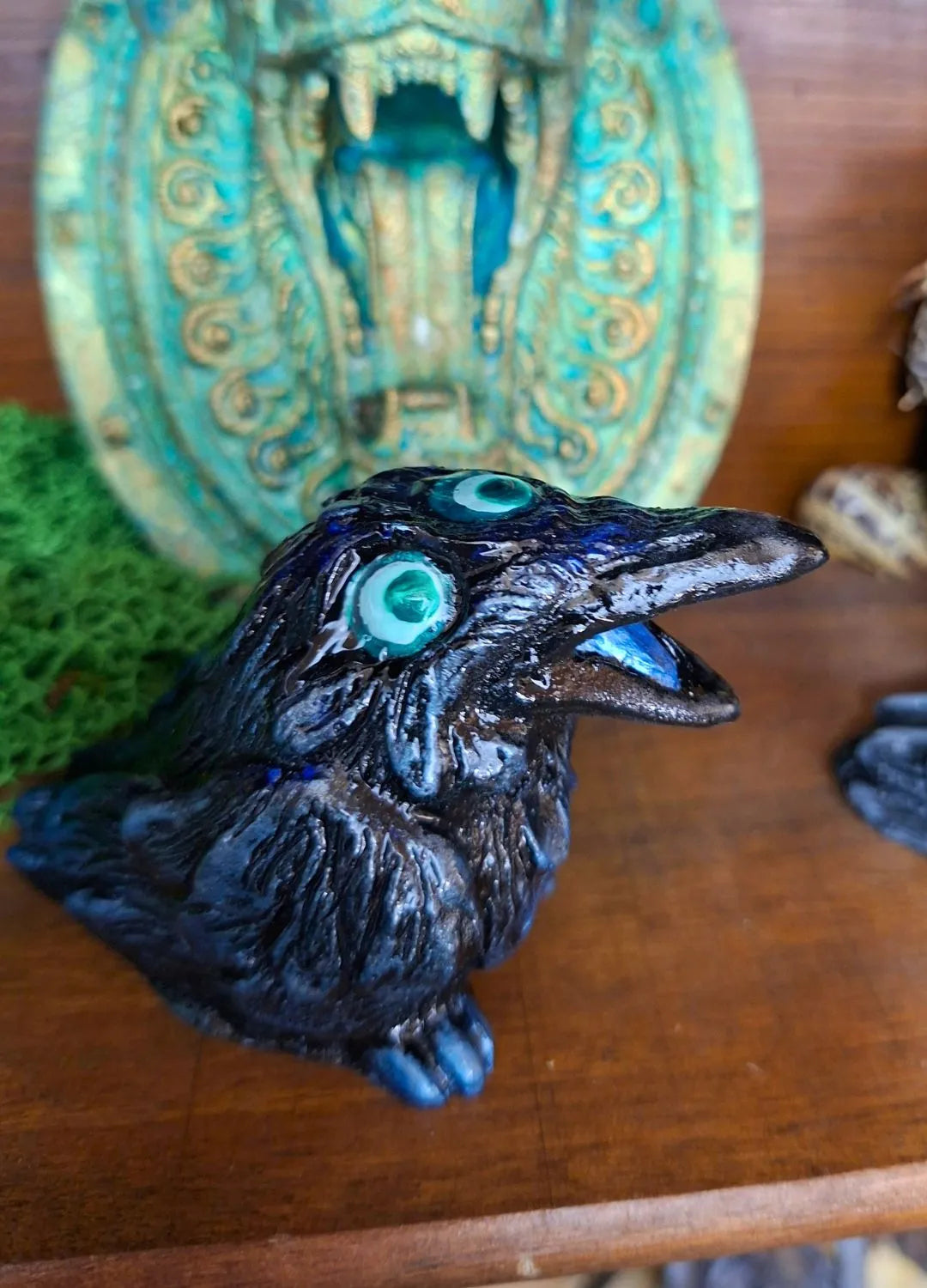 A dark black resin raven sculpture with subtle metallic sheen, bright light-blue eyes, and an open beak, showcased against a fantasy backdrop.