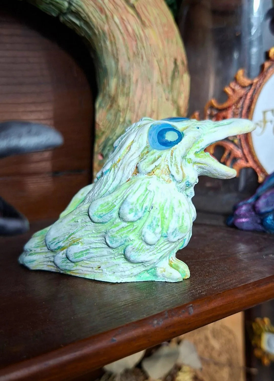 A fantasy raven sculpture made of resin with a pale green weathered finish and glowing blue eyes, perfect for dark academia bookshelf decor.