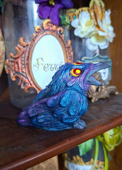 Handcrafted resin raven statue with a metallic deep purple and blue shifting finish and glowing yellow eyes on a dark wood cabinet.