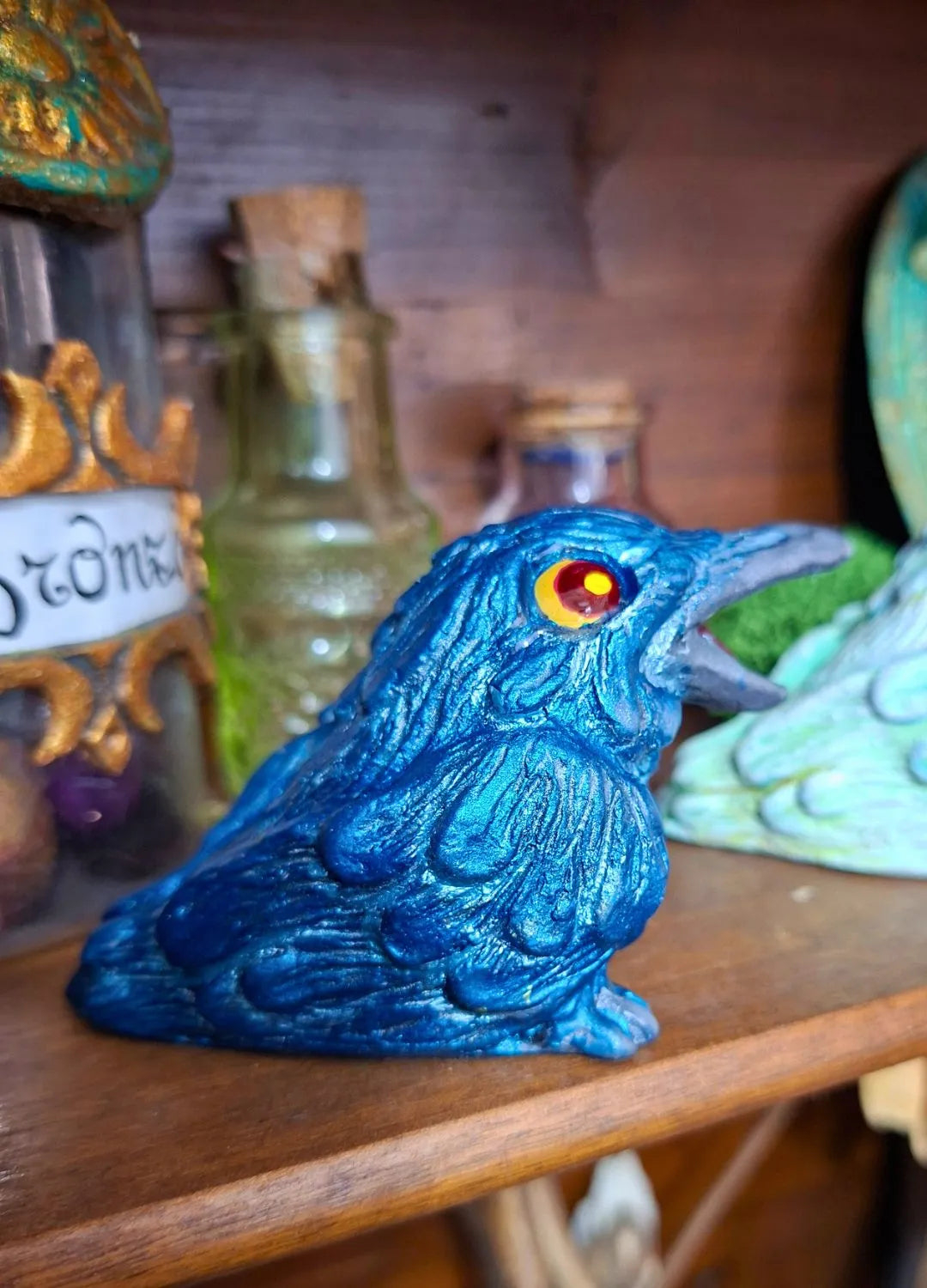A vibrant cobalt blue handmade raven figurine with detailed feathered textures and bright orange eyes, displayed in a curiosity cabinet setting.